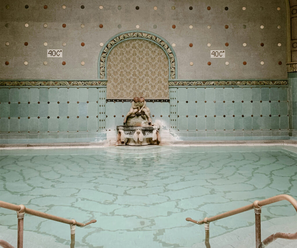 A imagem mostra uma piscina para banho turco com decoração clássica e elementos ornamentados. A piscina é cercada por azulejos verdes e brancos, com um chafariz central adornado com esculturas. A parede de fundo apresenta um arco decorado com padrões intrincados e duas placas indicando a temperatura de 40°C. O ambiente é caracterizado por uma paleta de cores verde e branca, com elementos de cerâmica e pedra.
