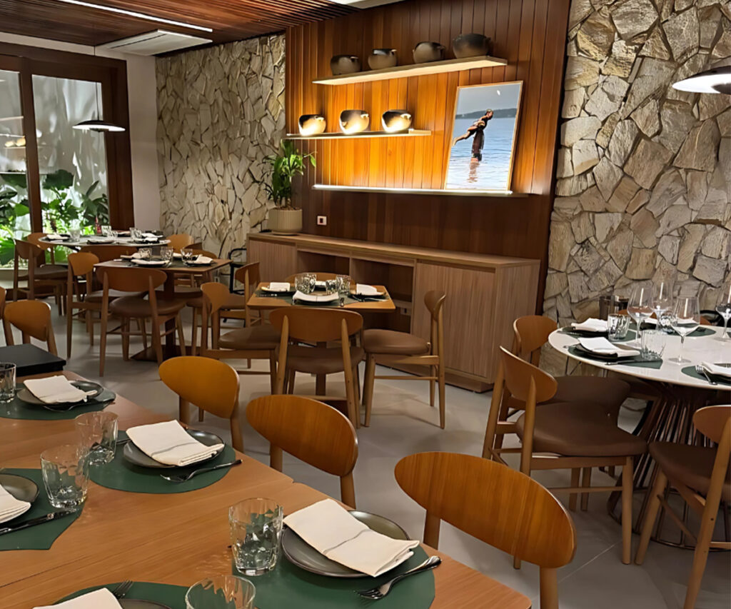 A imagem mostra o interior do restaurante Banzeiro. Moderno com decoração de interiores brasileira. O ambiente é composto por mesas de madeira com cadeiras combinando, todas arrumadas com talheres e copos. As paredes são revestidas com pedra natural e painéis de madeira, criando uma atmosfera quente e acolhedora. Uma prateleira com vasos e uma tela com uma imagem de uma pessoa na praia adicionam um toque artístico ao espaço. A iluminação é suave e ambiente, contribuindo para a atmosfera convidativa.