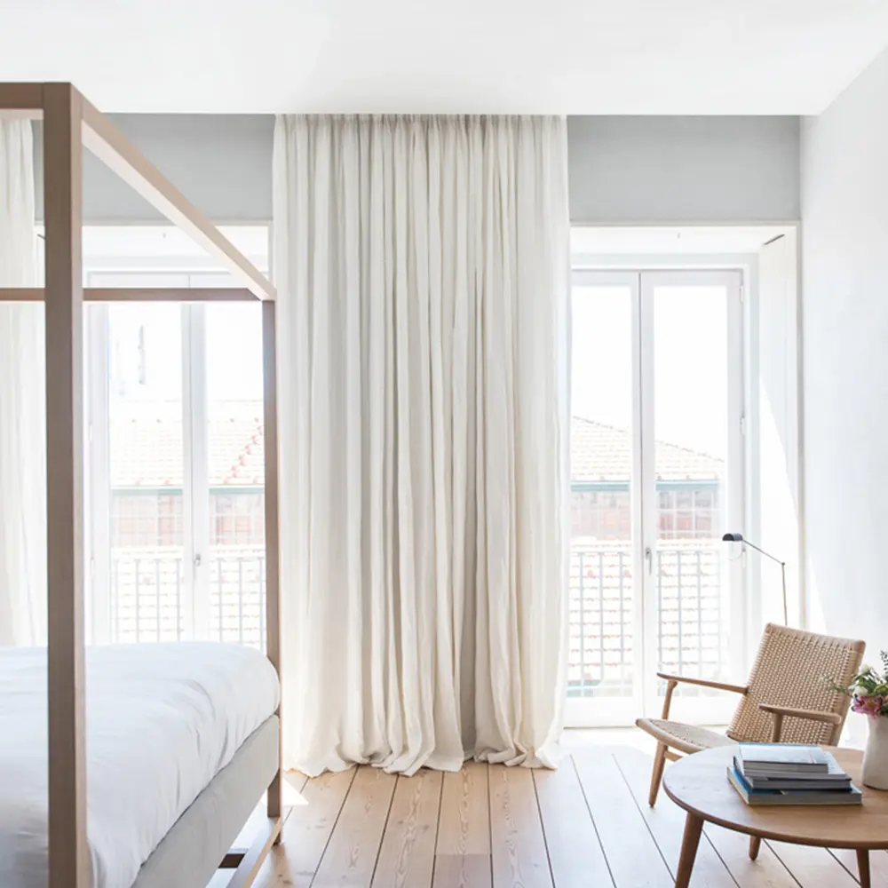 A imagem mostra um quarto moderno com uma cama de dossel de madeira, lençóis brancos e uma varanda com vista para edifícios. O quarto possui um design minimalista com cores claras e elementos de madeira. Há uma poltrona de vime ao lado de uma mesa de centro com livros e uma planta.