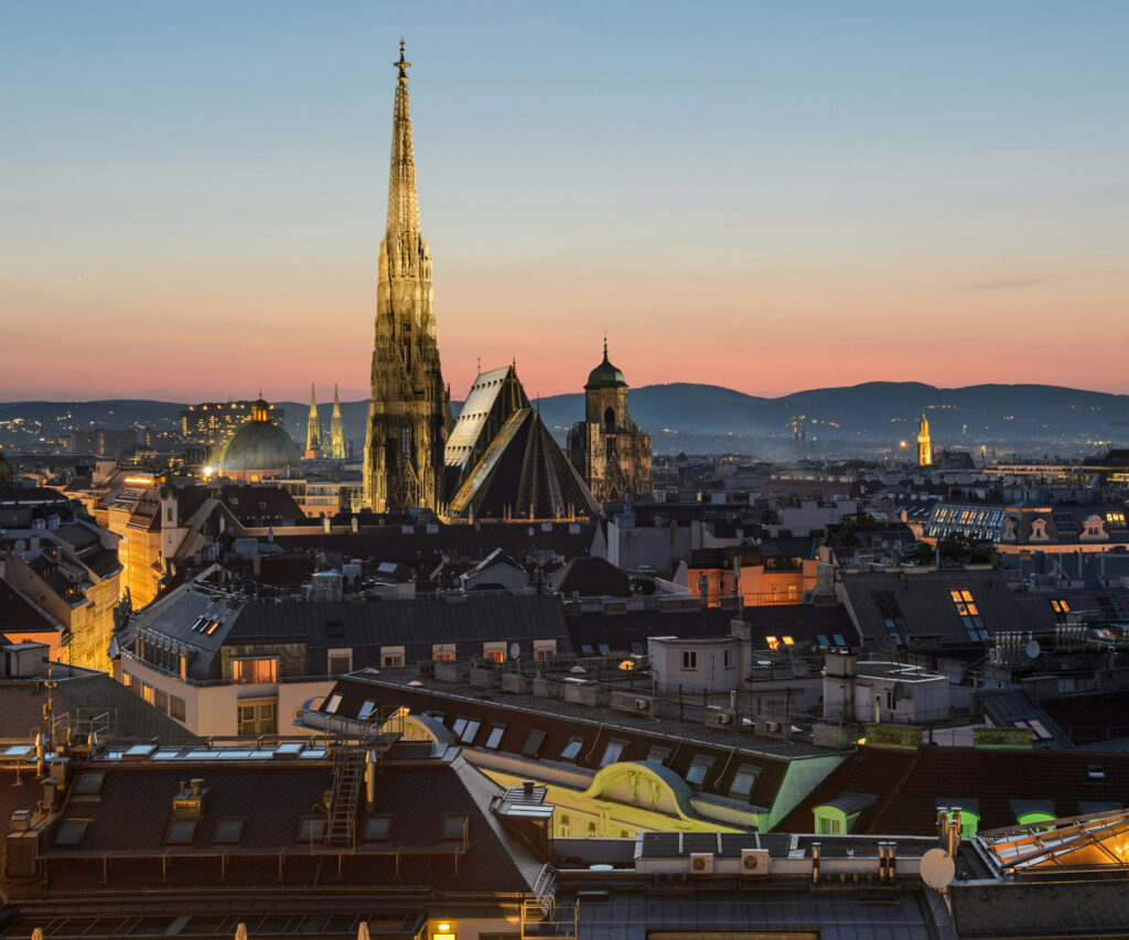 A imagem captura uma vista deslumbrante da paisagem urbana de Viena ao pôr do sol. A catedral, com suas torres imponentes e detalhes arquitetônicos intricados, destaca-se contra o céu azul claro. A cidade está banhada por uma luz quente e dourada, criando uma atmosfera serena e majestosa. Os edifícios ao redor da catedral são uma mistura de estilos arquitetônicos, com telhados de telhas e fachadas históricas. Ao fundo, as montanhas adicionam profundidade à cena, aumentando a beleza natural do cenário urbano.