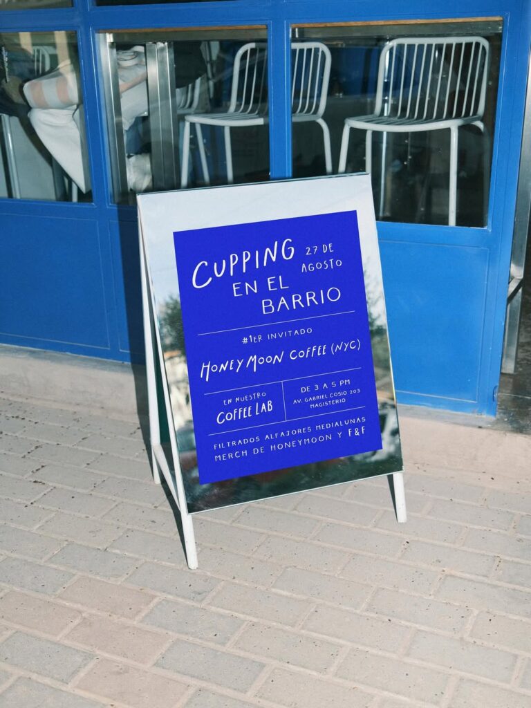 A imagem mostra uma placa de evento para um evento de degustação de café em um café moderno. A placa está em um suporte de metal e tem um fundo azul com texto branco. O café tem uma decoração minimalista com cadeiras de metal e uma fachada azul.