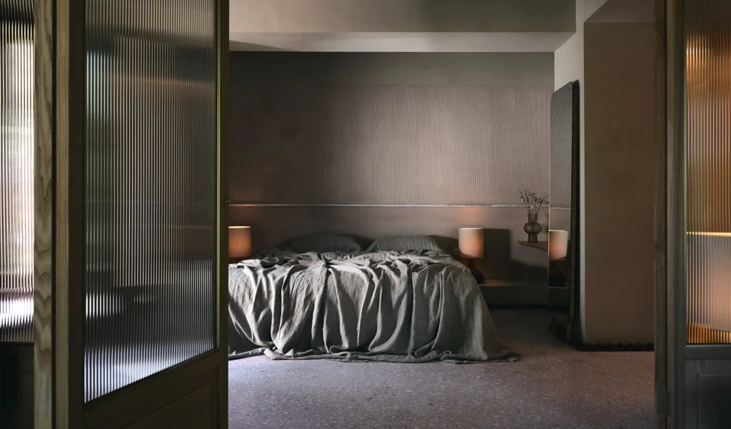 O CTRLZAK studio criou um quarto moderno e sofisticado com uma paleta de cores neutras. A cama está coberta com um edredom cinza e há duas mesas de cabeceira com lâmpadas. As paredes têm texturas e os materiais incluem madeira e tecido.