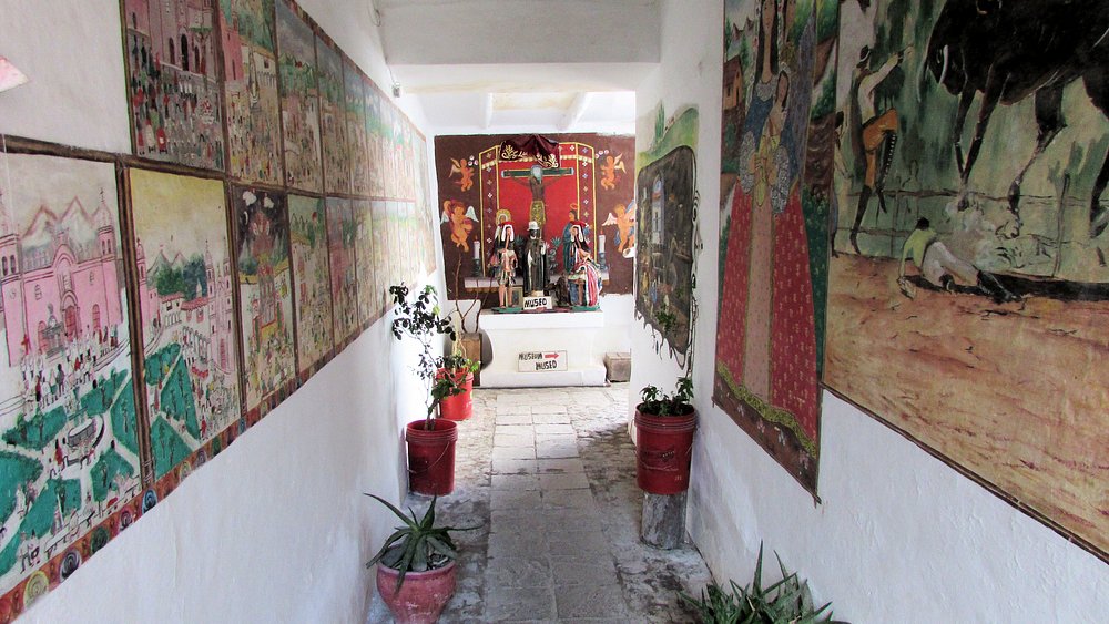 O corredor do Museu Hilario Mendivil apresenta murais coloridos com cenas de vida e cultura, plantas em vasos e uma decoração vibrante. As esculturas em madeira com longos pescoços inspiradas em lhamas são um destaque único.