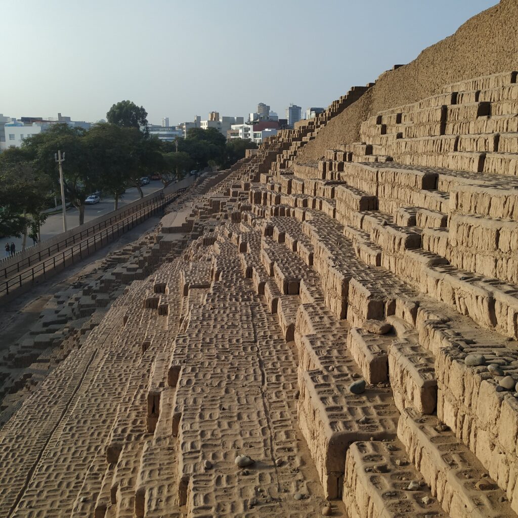 A imagem mostra a pirâmide Huaca Pucllana em Lima, Peru, uma estrutura arqueológica significativa feita de adobe e argila. A pirâmide é cercada por um ambiente urbano moderno, com árvores e edifícios ao fundo. A estrutura é composta por múltiplos níveis com escadas e recantos, destacando a arquitetura pré-hispânica.