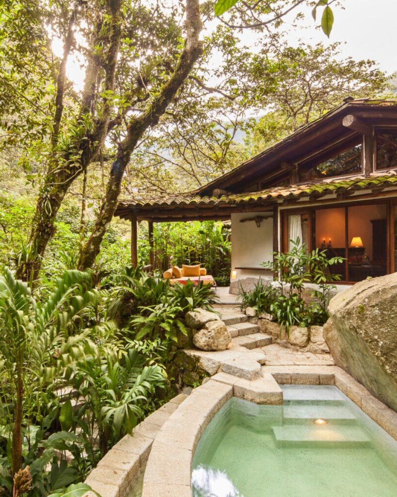 A imagem mostra uma casa tropical com uma área de estar ao ar livre cercada por vegetação exuberante. Há uma piscina pequena e retangular no primeiro plano, com degraus de pedra levando até ela. A casa tem um telhado de telhas e grandes janelas que permitem a entrada de luz natural. A área de estar ao ar livre inclui móveis confortáveis e plantas em vasos. A decoração é natural e rústica, com um foco em integração com a natureza.