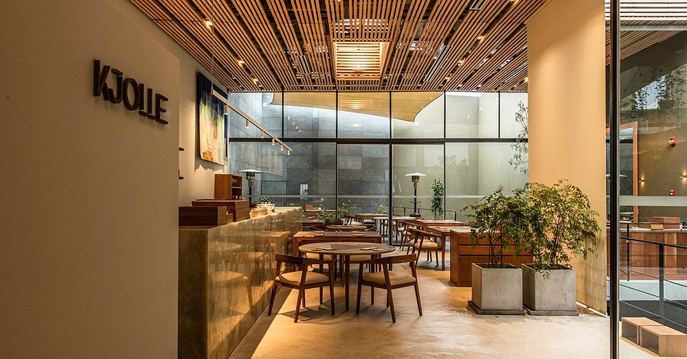 A imagem mostra o interior do Restaurante Mayta em Lima, no Peru, com uma decoração moderna e acolhedora. A paleta de cores predominantes inclui tons terrosos e neutros, como cinza e bege. Os móveis incluem mesas de pedra e sofás confortáveis com almofadas. Objetos decorativos como luminárias e xícaras de cerâmica estão estrategicamente colocados. O ambiente é realçado por uma vegetação exuberante ao fundo, criando uma atmosfera serena e sofisticada.