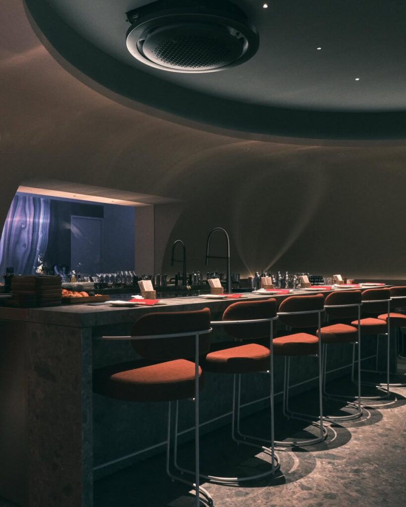 O interior do restaurante LadyBee apresenta um ambiente intimista com uma paleta de cores escuras e acolhedoras. O bar é equipado com várias garrafas e copos, e há várias banquetas vermelhas com assentos acolchoados. O design é moderno, com elementos de iluminação suave e materiais como metal e tecido.