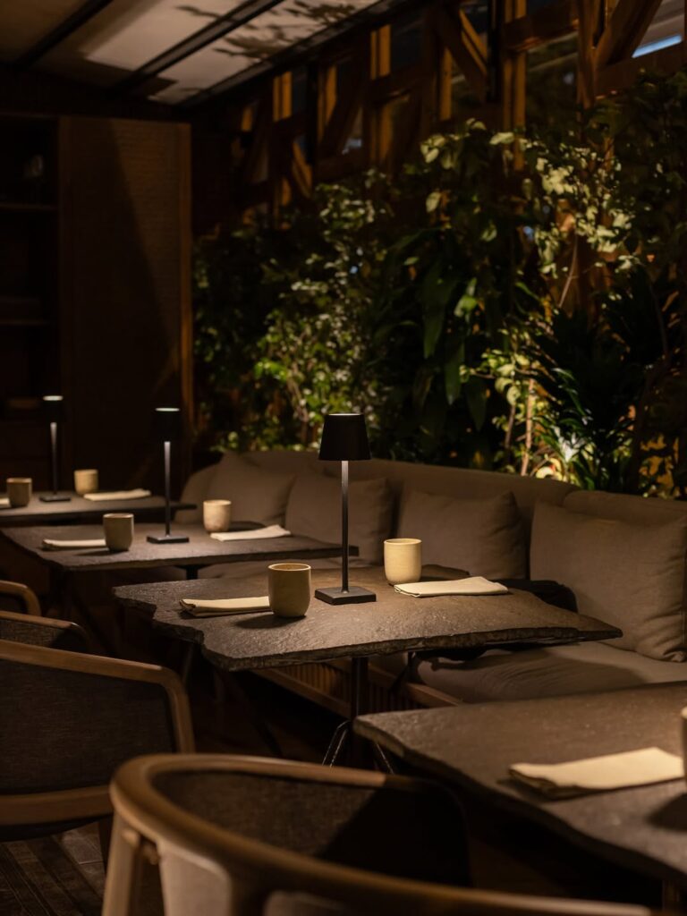 A imagem mostra o interior do Restaurante Mayta em Lima, no Peru, com uma decoração moderna e acolhedora. A paleta de cores predominantes inclui tons terrosos e neutros, como cinza e bege. Os móveis incluem mesas de pedra e sofás confortáveis com almofadas. Objetos decorativos como luminárias e xícaras de cerâmica estão estrategicamente colocados. O ambiente é realçado por uma vegetação exuberante ao fundo, criando uma atmosfera serena e sofisticada.