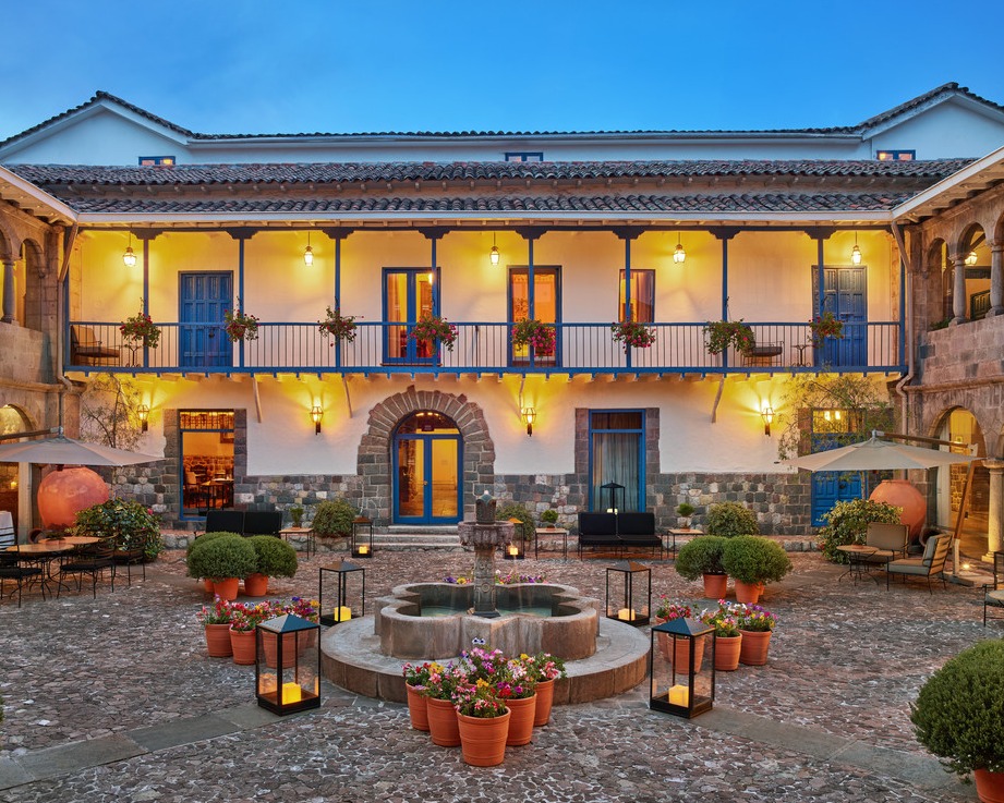 A imagem retrata o pátio interno do Palácio del Inka, um hotel de luxo em Cusco. O pátio é adornado com plantas em vasos, lanternas e um chafariz central. A arquitetura colonial é destacada por paredes brancas, janelas azuis e varandas com flores. O ambiente é acolhedor e sofisticado, com iluminação suave e decoração cuidadosa.