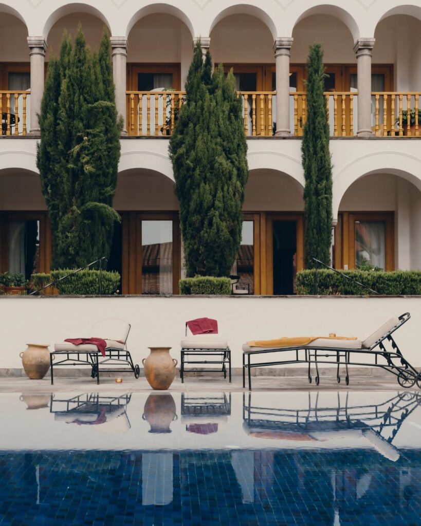 A imagem mostra uma área de piscina externa no Palácio Nazarenas, um local de luxo em Cusco. A piscina é cercada por espreguiçadeiras com almofadas coloridas e grandes vasos de plantas. Ao fundo, há um edifício com colunas e varandas de madeira, proporcionando uma atmosfera tranquila e sofisticada.