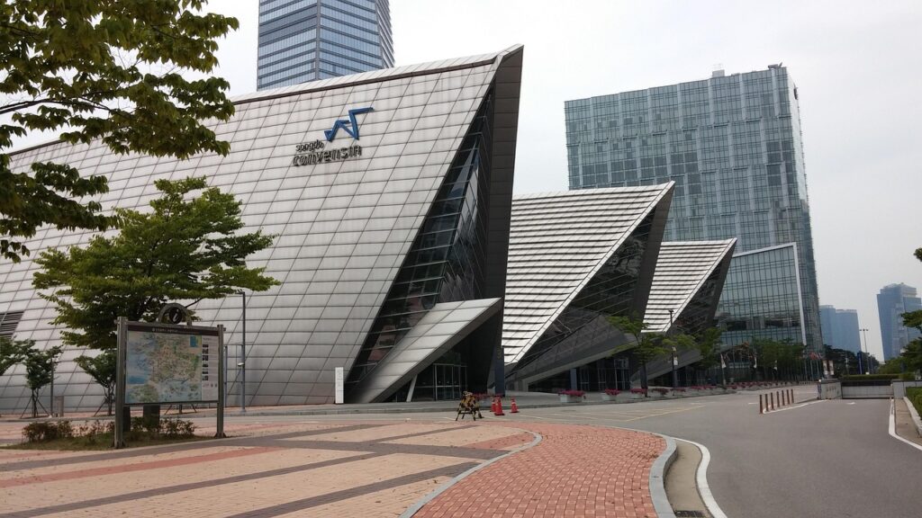 A imagem mostra um edifício moderno com um design arquitetônico único em forma de triângulo, localizado em uma cidade high tech. O edifício tem uma fachada de vidro e aço, com um design angular distintivo. Ao fundo, há arranha-céus modernos e árvores que adicionam um toque de verde ao ambiente urbano. A área ao redor é pavimentada com tijolos e há uma placa com um mapa na frente do edifício. Ainda assim, é uma cidade vazia.