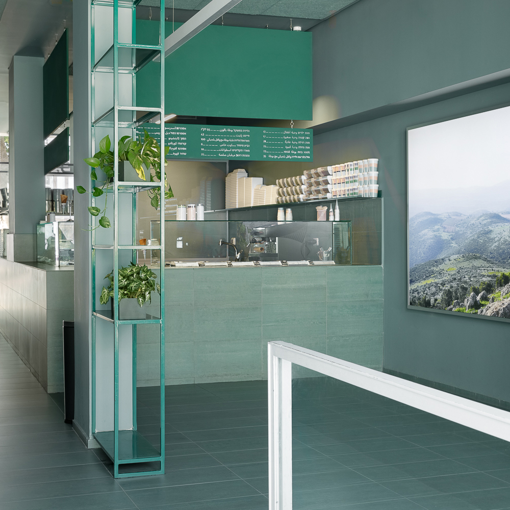 Decoração minimalista. A sorveteria Buza em Tel Aviv apresenta um design minimalista com uma paleta de cores que combina tons de cinza e verde azulado. O ambiente é caracterizado por móveis de vidro e elementos de metal, criando um espaço que dialoga com a arquitetura industrial ao redor. Prateleiras de vidro com plantas, balcões de atendimento e uma grande imagem de paisagem contribuem para a estética moderna e clean.