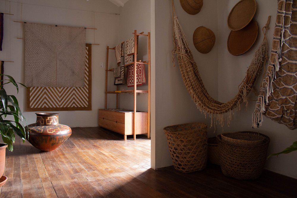 O espaço da Xapiri Ground apresenta uma decoração rústica que reflete a riqueza do artesanato da região. A imagem destaca um ambiente com móveis de madeira, tapetes tecidos à mão, cestas de vime e vasos de cerâmica. As paredes são adornadas com tecidos e cestas, criando uma atmosfera acolhedora e autêntica. A paleta de cores é predominantemente terrosa, com tons de marrom, bege e branco. O estilo de decoração é boho-chic, combinando elementos naturais e artesanais. O ambiente parece ser uma sala de estar ou um espaço de convivência, ideal para relaxamento e apreciação da cultura local.