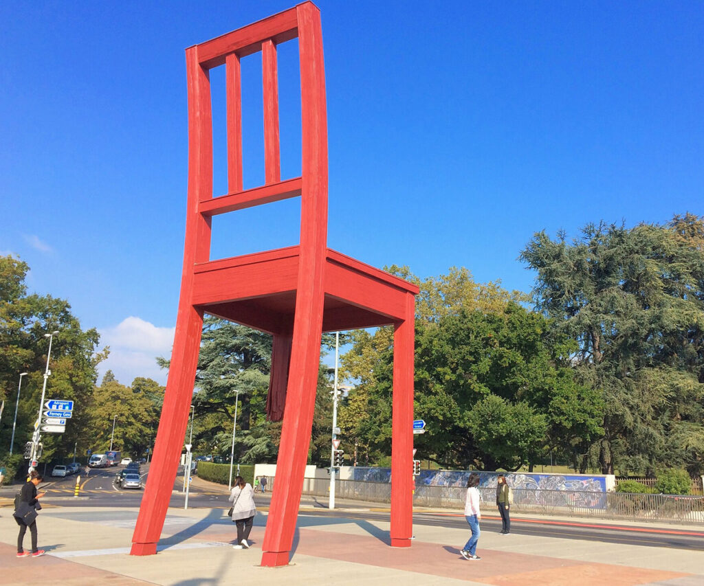 Genebra Suíça: grande escultura de cadeira vermelha em praça pública com pessoas caminhando.