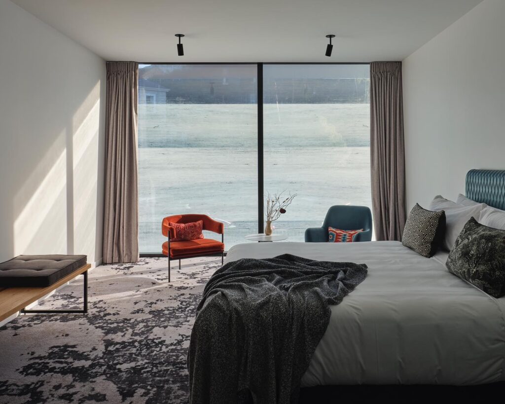 Hotel EBB Dunedin: quarto moderno com vista panorâmica, cama elegante e poltrona vermelha junto a janela ampla
