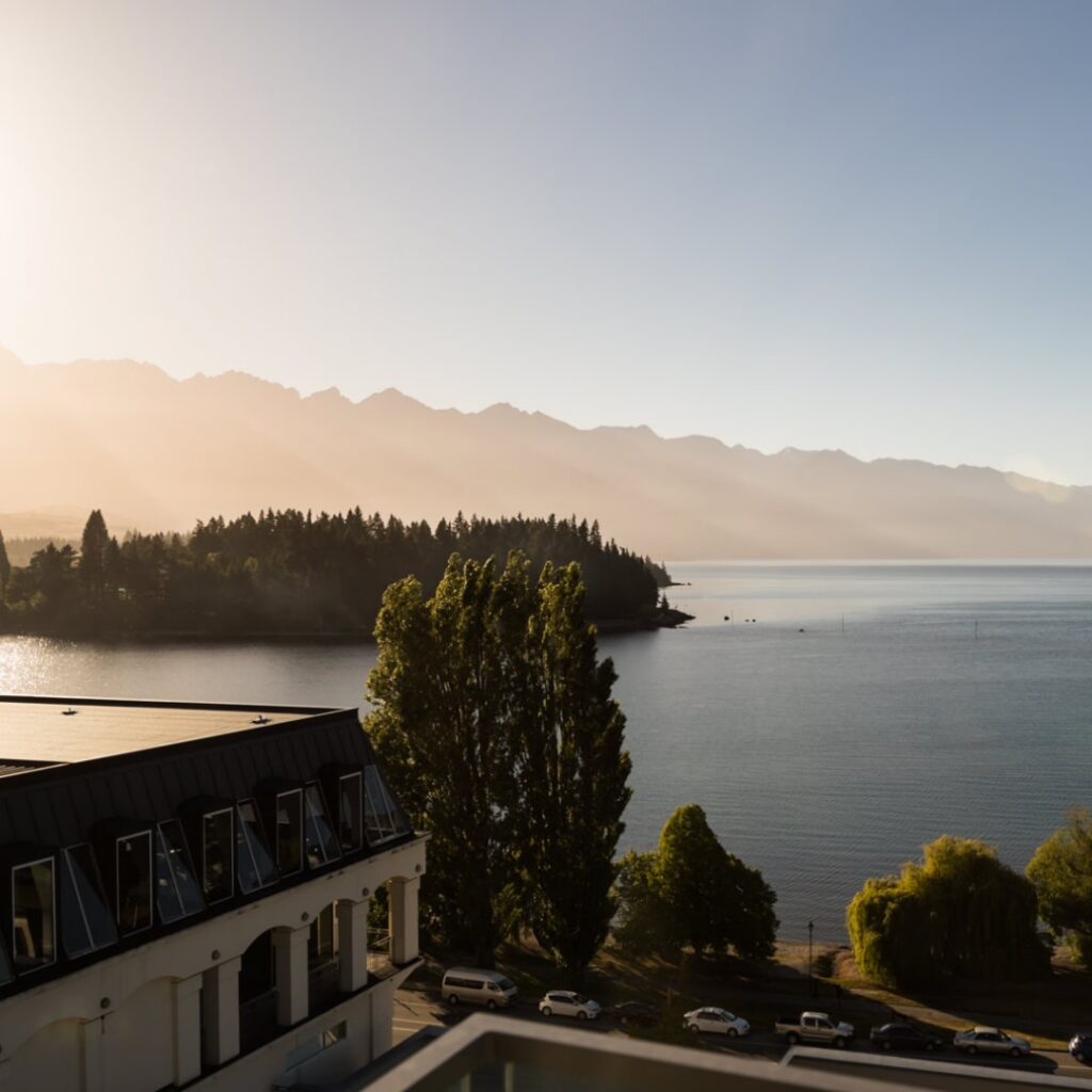 Viagem para Nova Zelândia: vista luxuosa de lago com quarto moderno, sofá e mesa com livro, paisagem de montanhas e água cristalina