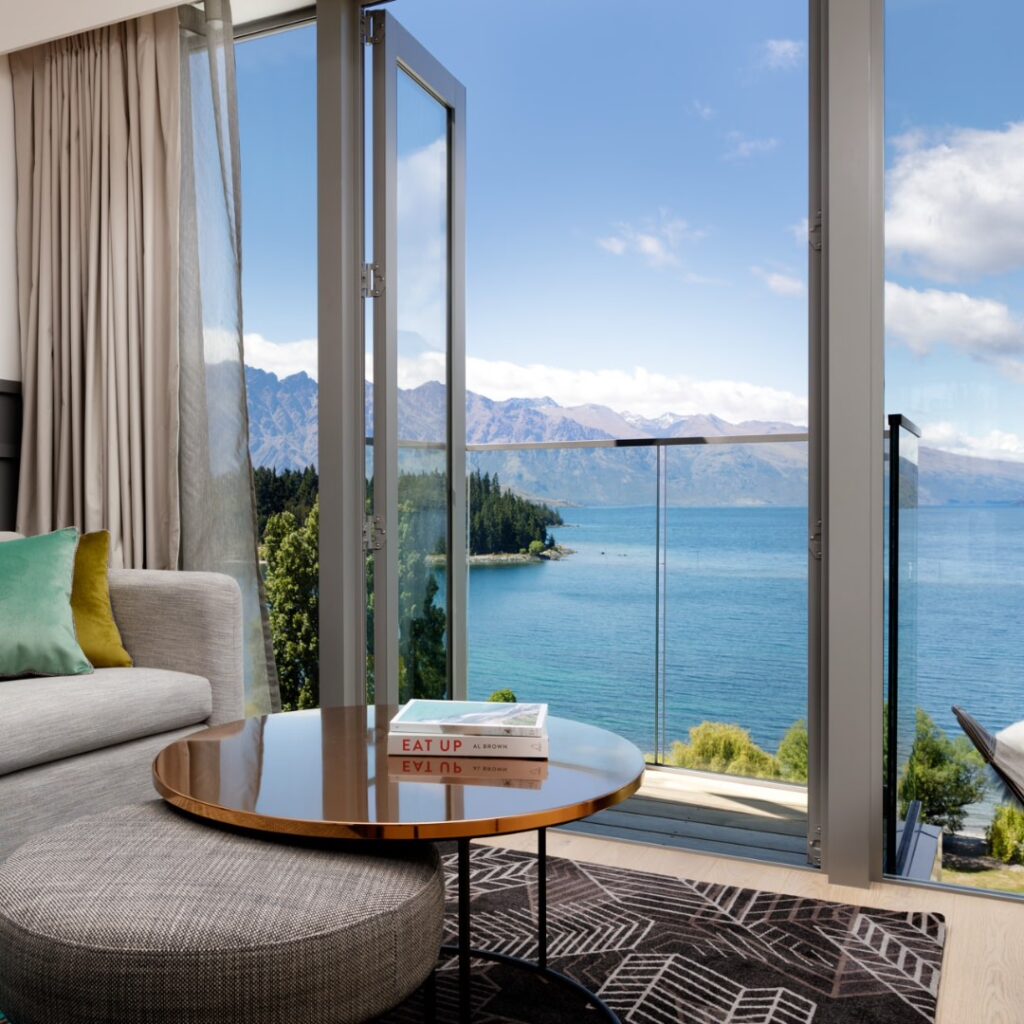Viagem para Nova Zelândia: vista luxuosa de lago com quarto moderno, sofá e mesa com livro, paisagem de montanhas e água cristalina