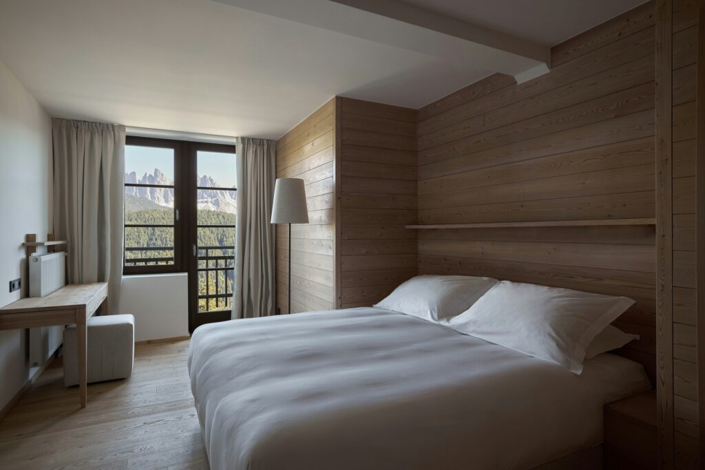 Quarto de hotel italiano com paisagem dos Dolomitas enquadrada por janela de esquadria preta, revestimento total em madeira de abeto.