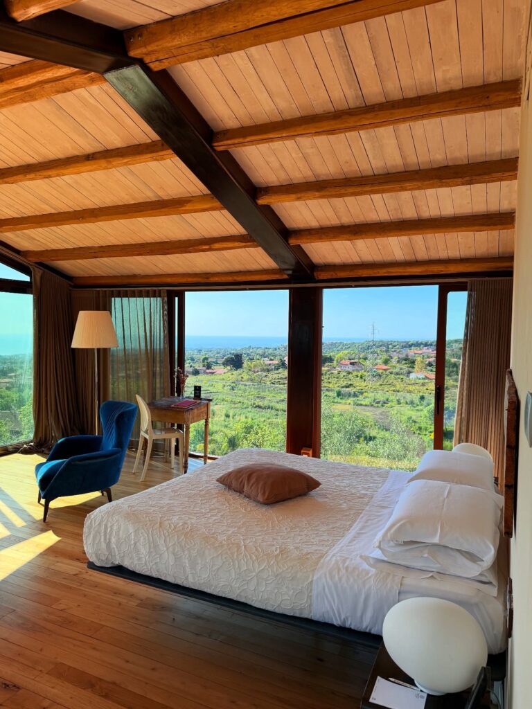 Quarto de hotel italiano com paisagem campestre vista através de grandes janelas, destacando teto de madeira e poltrona azul de design.