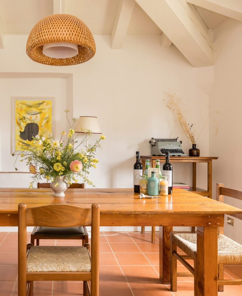 Sala de jantar de hotel italiano com paisagem toscana, mesa de madeira rústica, piso cotto terracota e pendente de vime artesanal.