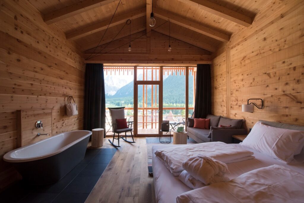 Suíte de hotel italiano com paisagem dos Dolomitas, banheira autônoma cinza, teto de duas águas e paredes em pinho zirbel.