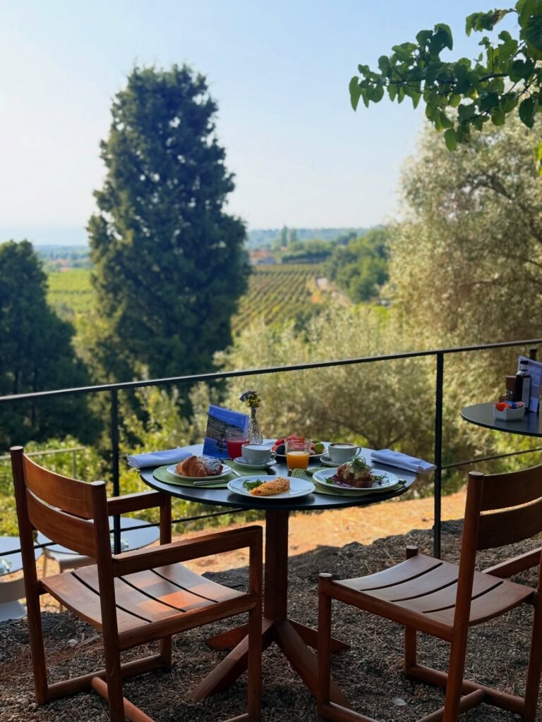 Mesa de café da manhã em terraço ao ar livre de hotel italiano com paisagem de vinhedos e oliveiras ao fundo.