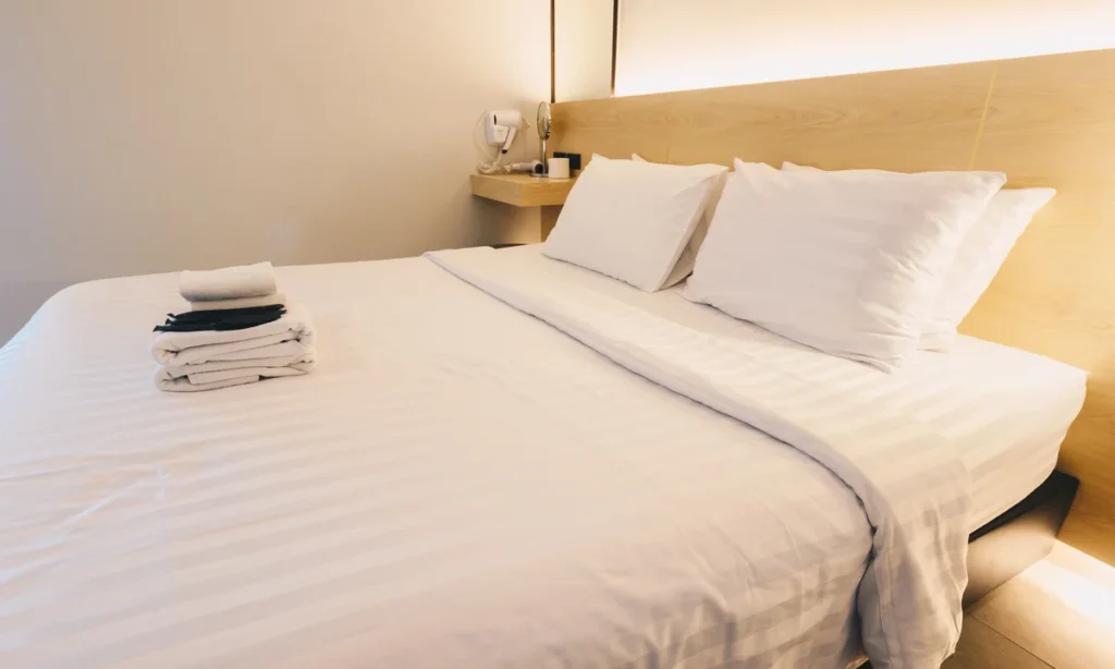 Cama de hotel minimalista com enxoval branco e iluminação embutida, destacando as novas regras check in e check out e padrões de higiene.
