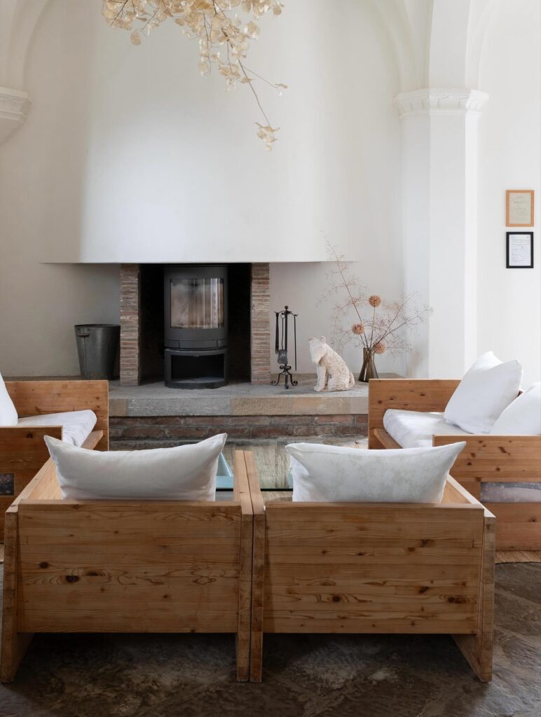 Sala com lareira histórica de hotel italiano com paisagem, poltronas de madeira bruta e mobile de plantas secas.