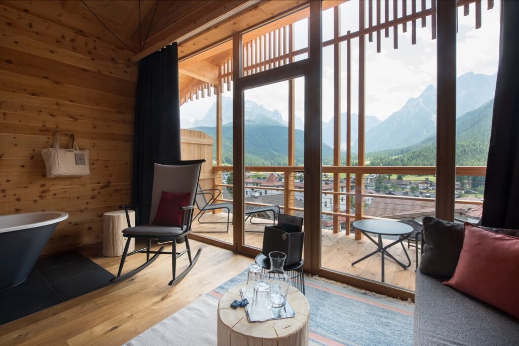 Varanda de hotel italiano com paisagem dos Dolomitas e vale de Sexten, enquadrada por caixilho de madeira e cadeira de balanço.