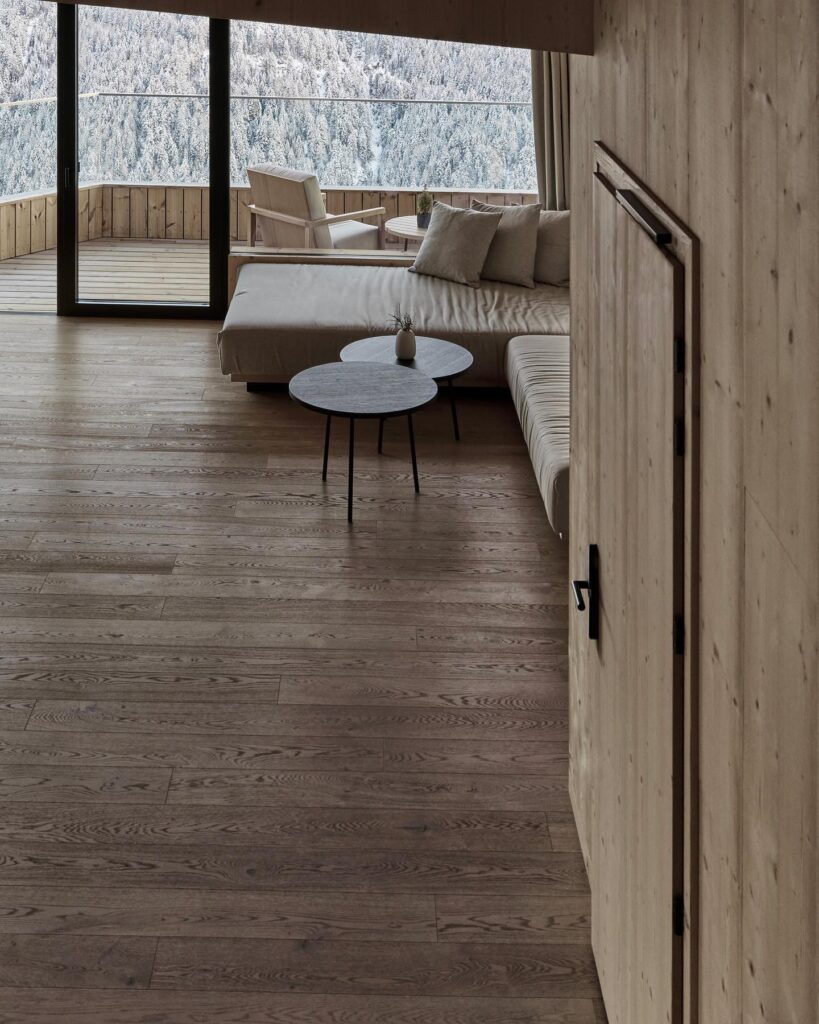 Sala de estar de hotel italiano com paisagem de floresta nevada, revestimento total em madeira de abeto e varanda com vista.