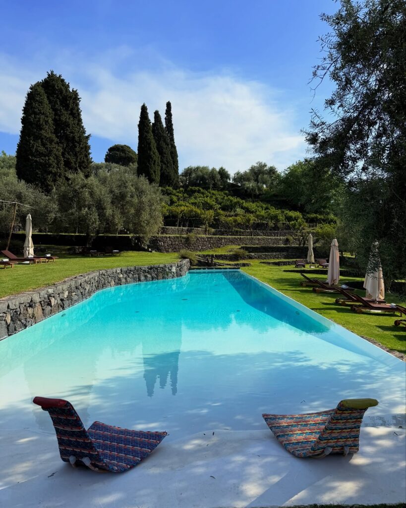 Piscina de design em hotel italiano com paisagem de colinas sicilianas, muros de pedra e espreguiçadeiras coloridas.