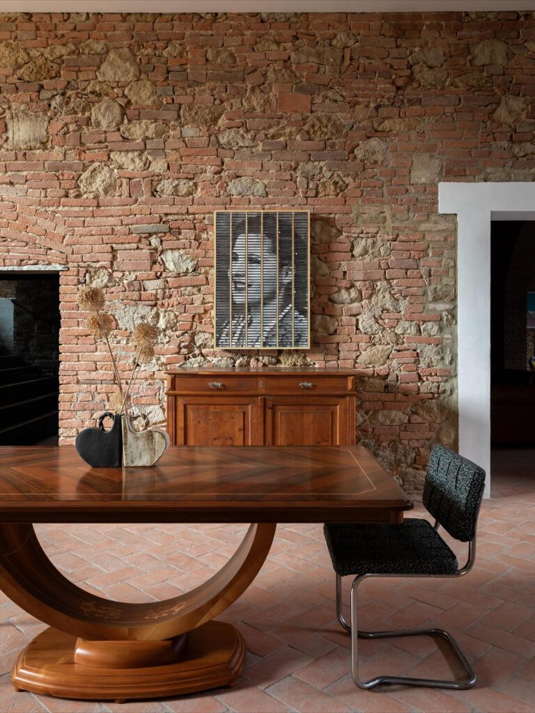 Ambiente de hotel italiano com paisagem e tijolo histórico exposto, mesa art déco escultórica e obra de arte contemporânea.