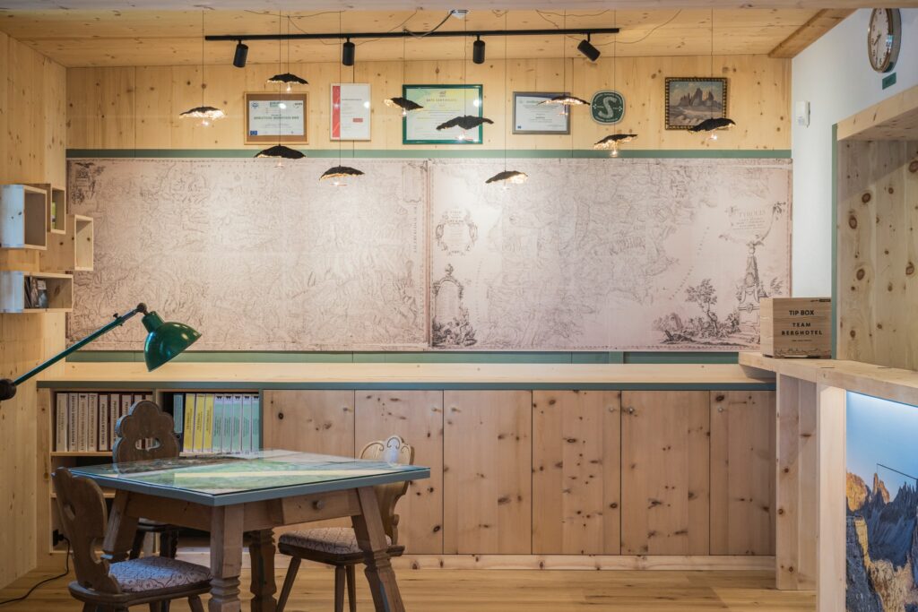 Recepção de hotel italiano com paisagem alpina, parede com mapa histórico dos Dolomitas, balcão em pinho zirbel e mesa tirolesa.