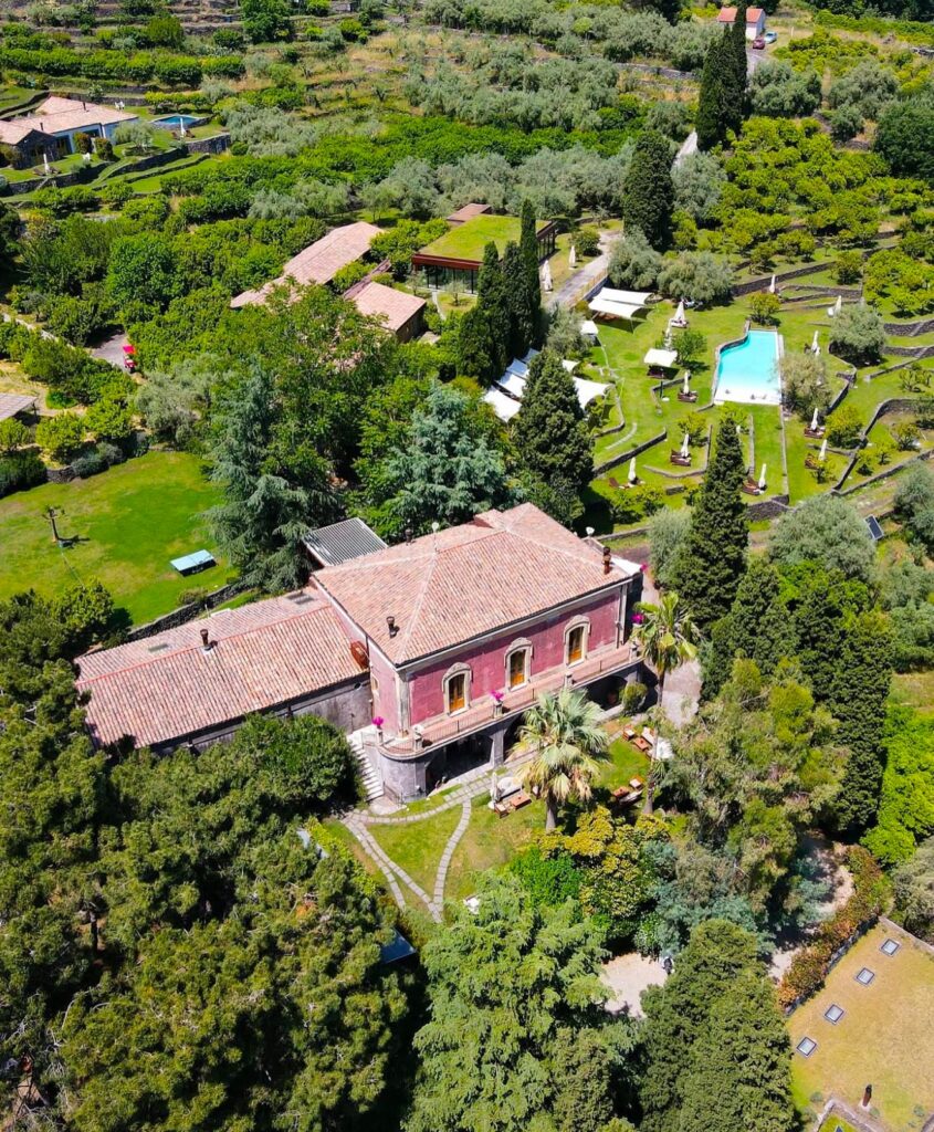 Vista aérea de hotel italiano com paisagem siciliana, destacando prédio histórico terracota, piscina e jardins em socalcos.