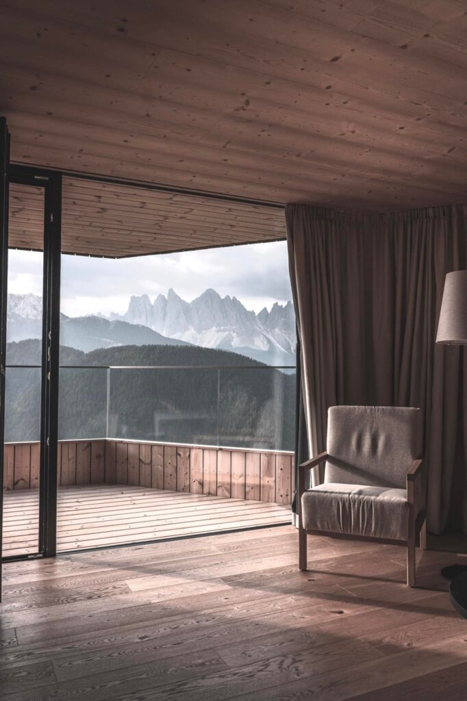 Poltrona em quarto de hotel italiano com paisagem dos Dolomitas através de janela de canto, varanda de madeira e vista para os picos.