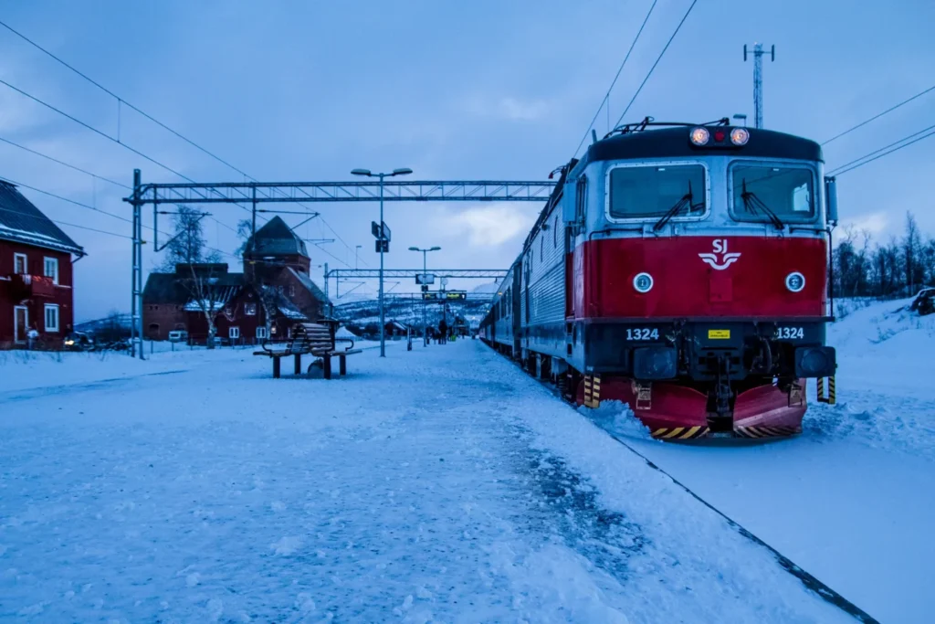 Locomotiva vermelha e prata da SJ número 1324 parada em plataforma coberta de neve na rota do trem da aurora boreal entre Estocolmo e Narvik