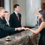 Recepcionista entregando cart&atilde;o de acesso &agrave; h&oacute;spede em balc&atilde;o de m&aacute;rmore, sob as novas regras para check in e check out.