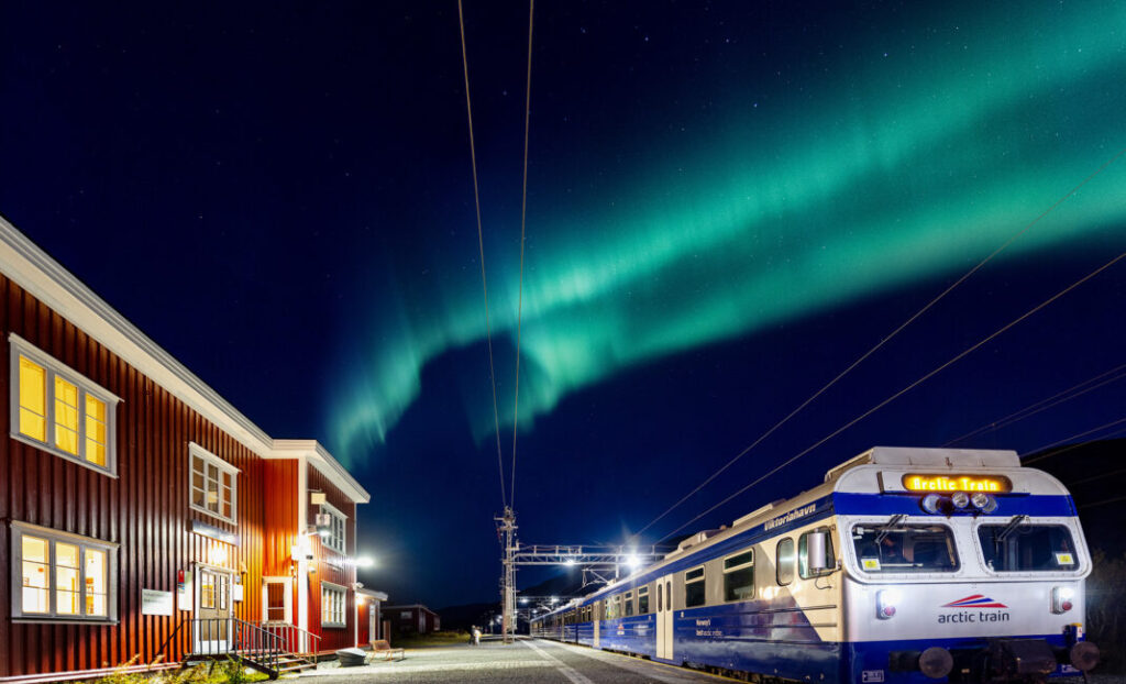 Trem da aurora boreal Arctic Train parado na estação de Katterat à noite com aurora boreal verde no céu e casarão histórico iluminado ao fundo