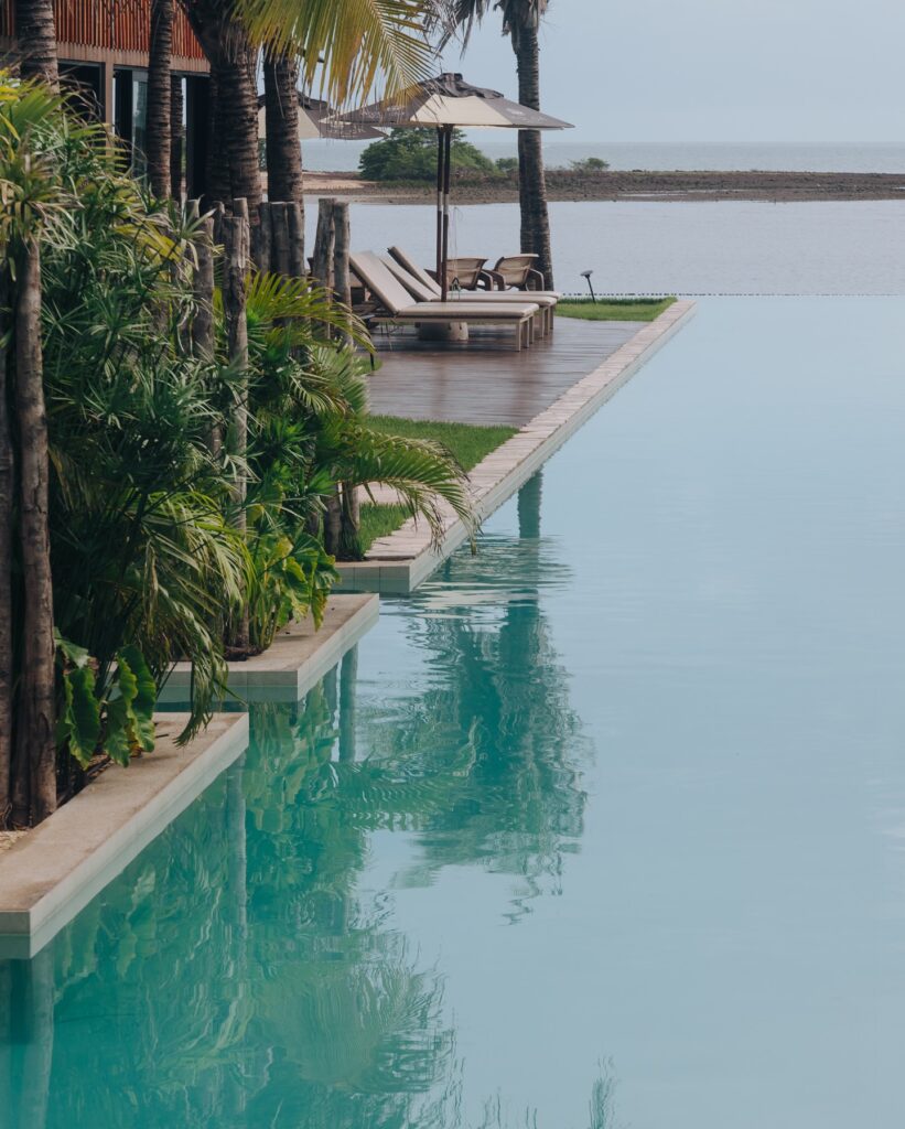Piscina de borda infinita rodeada por palmeiras e deck de madeira, oferecendo um refúgio para páscoa tranquilo à beira-mar.