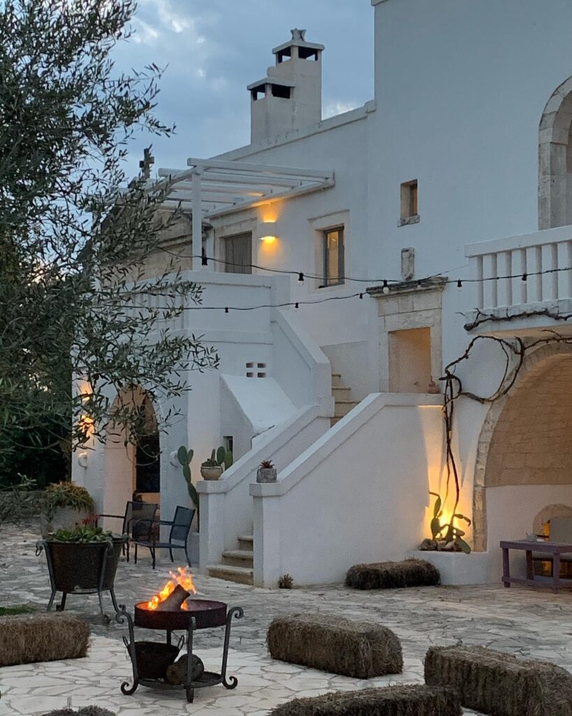 Fachada iluminada de hotel italiano com paisagem e lareira externa ao anoitecer em pátio de pedra.