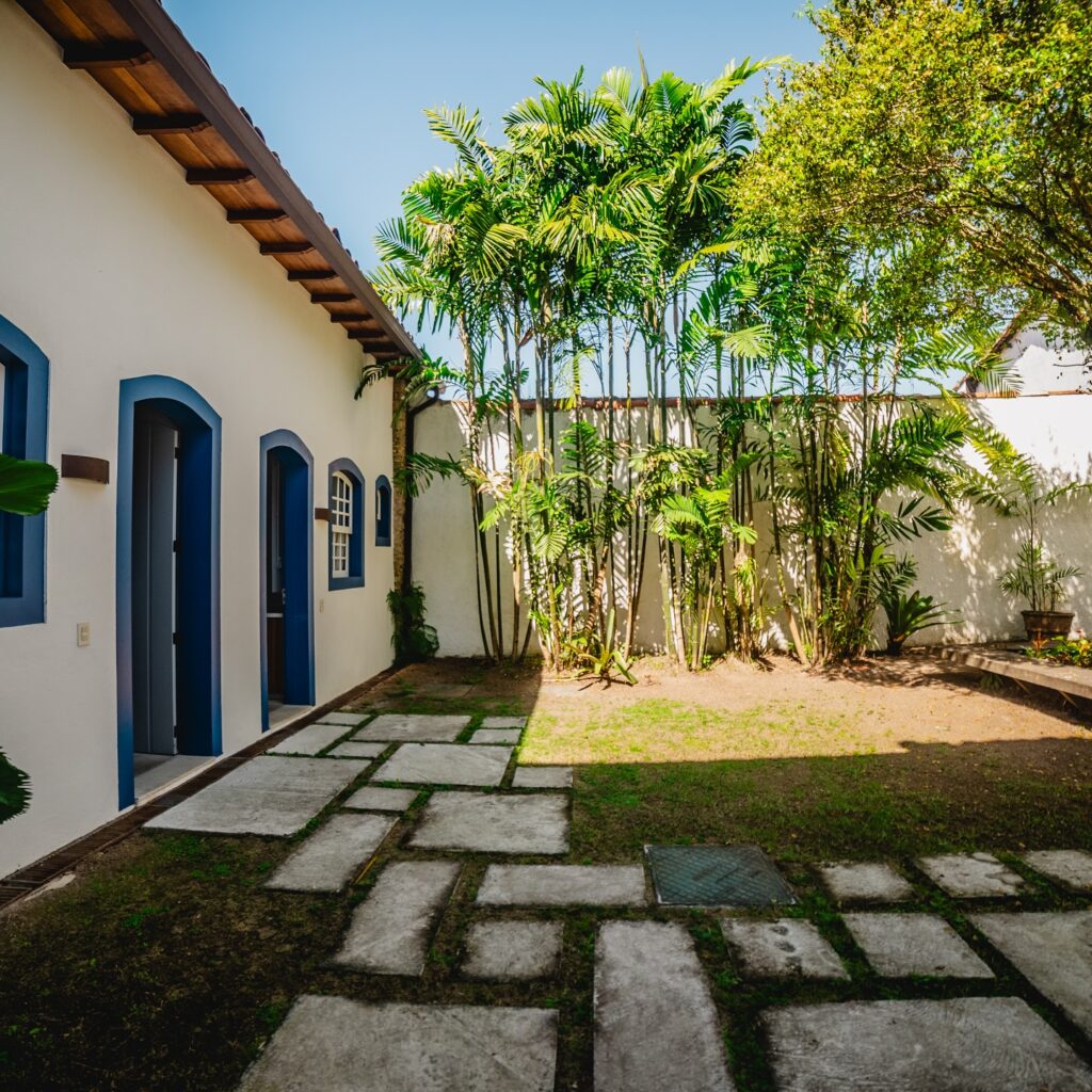 Pátio externo da Pousada Literária com arquitetura colonial branca e azul e paisagismo tropical sob luz solar direta.