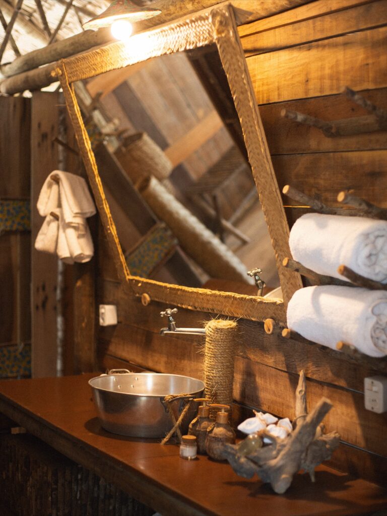 Banheiro rústico com cuba de metal, espelho de palha e detalhes em madeira no Rancho do Peixe.