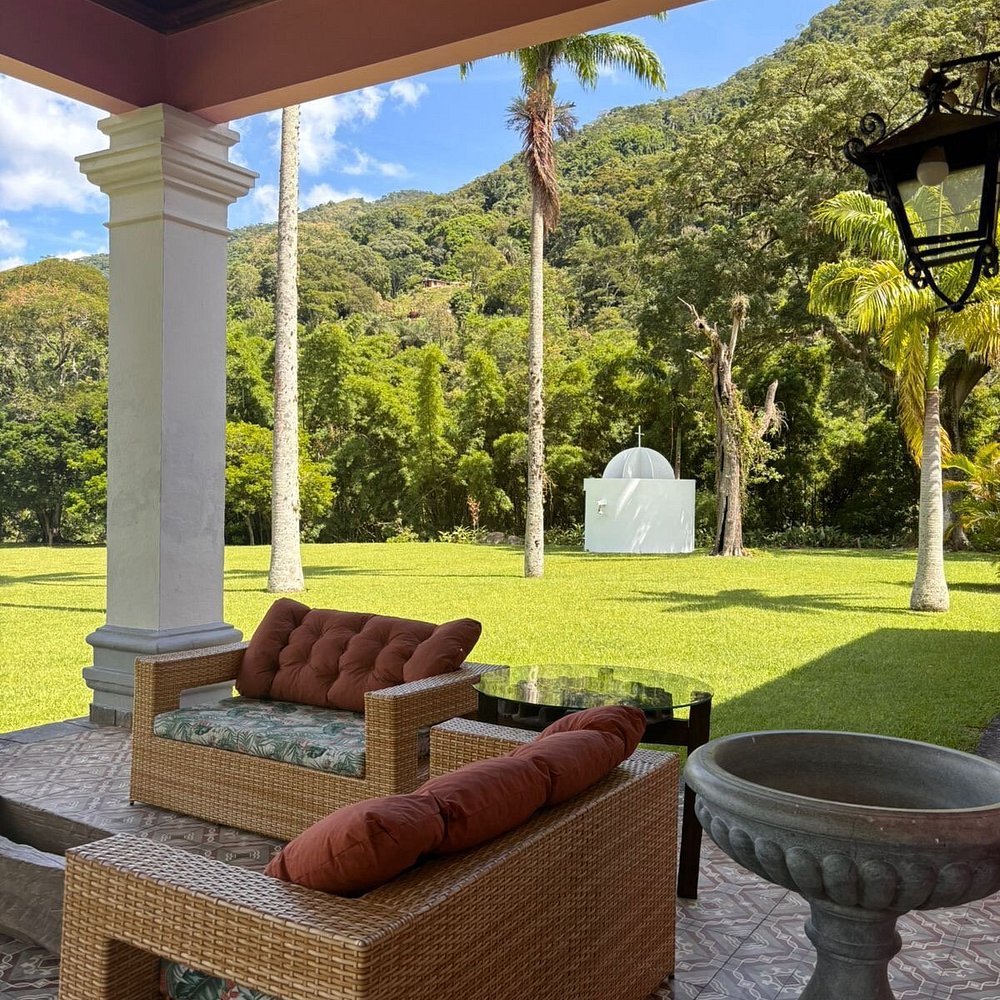 Mobiliário de vime em varanda com vista para jardim e capela modernista em turismo histórico de alto padrão.