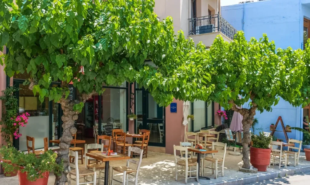 Terraço sombreado de café tradicional na Ilha de Creta com cadeiras de madeira e palha em ambiente acolhedor ao ar livre.
