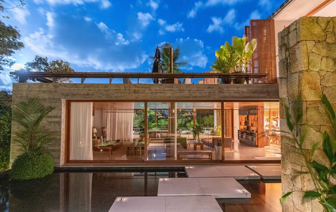 Iluminação noturna de casa de luxo para alugar em Itacaré com arquitetura moderna de vidro, concreto e espelho d'água iluminado.