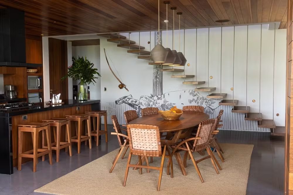 Sala de jantar com mesa de madeira e escada flutuante em casa de luxo para alugar em Itacaré, com design de interiores tropical moderno.