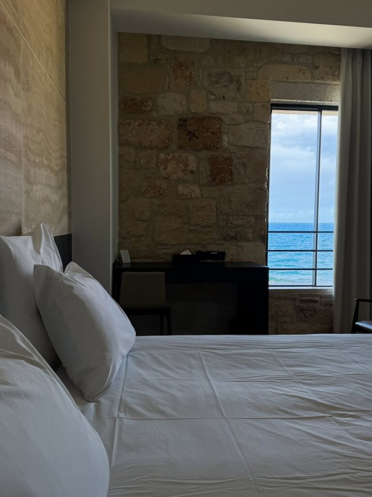 Quarto de luxo em um dos melhores hoteis da ilha de creta, destacando parede de pedra rústica e janela com vista para o mar azul.