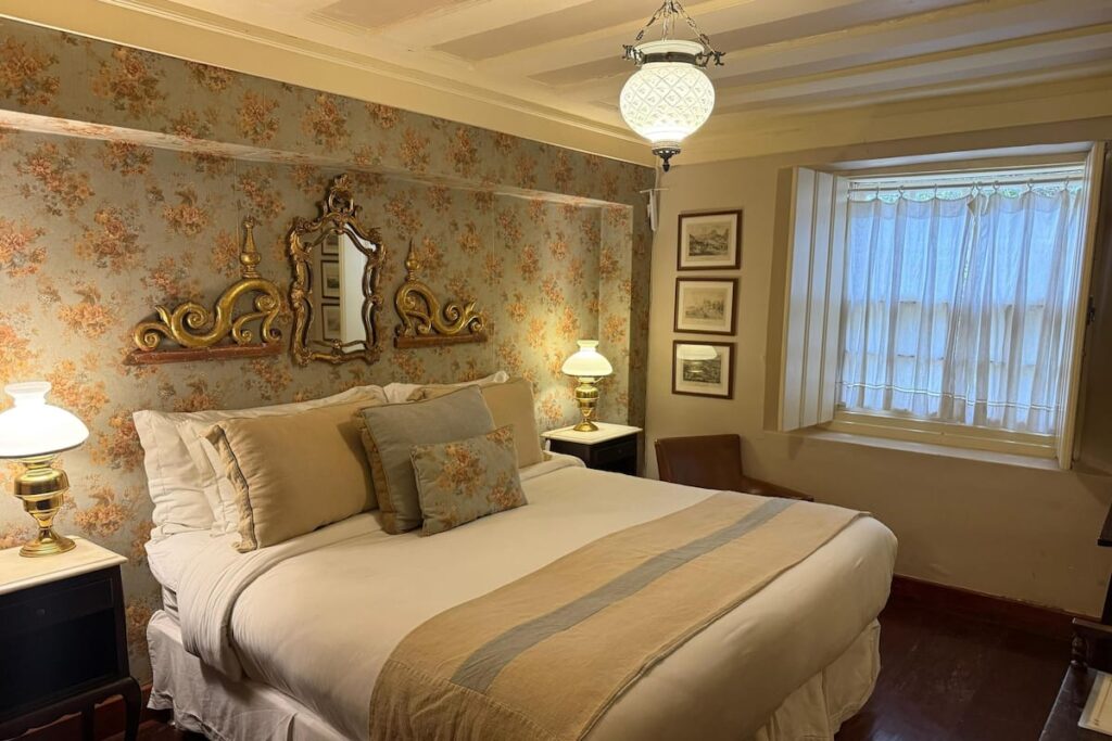 Quarto romântico com papel de parede floral e espelho veneziano em uma das elegantes fazendas históricas para se hospedar.