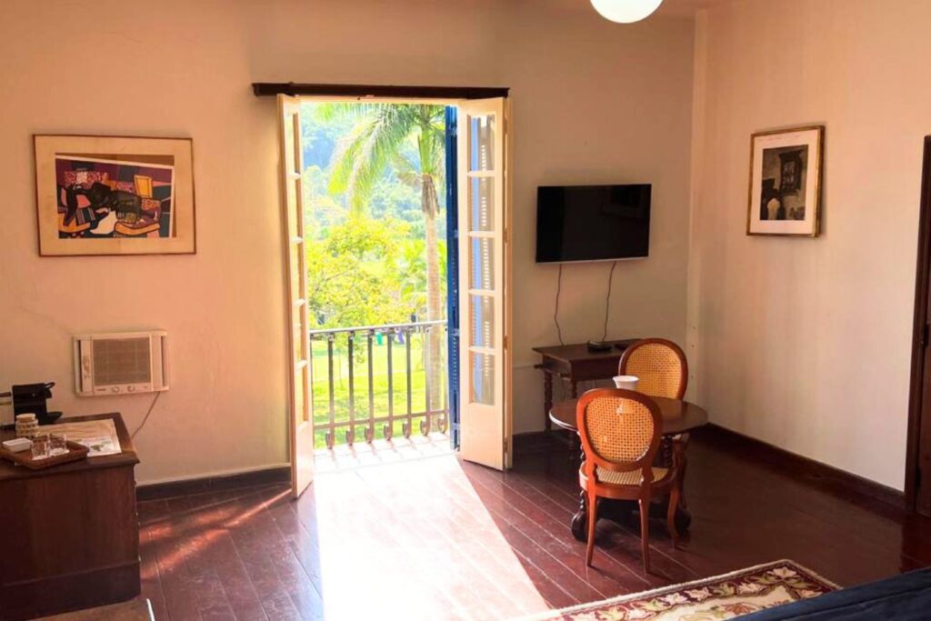 Quarto clássico com porta aberta para varanda em uma das fazendas históricas para se hospedar, com móveis de madeira e palhinha.