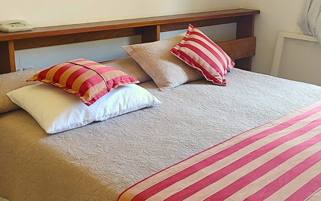 Cama confortável com cabeceira de madeira e almofadas listradas, detalhe em fazendas históricas para se hospedar.